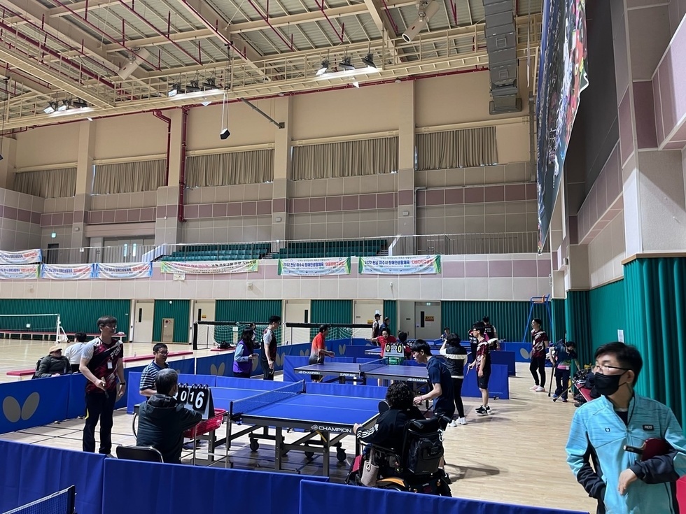 실내 체육관에서 20여명의 장애인과 비장애인이 함께 어울려 탁구 경기를 진행 중인 모습. 각 탁구대마다 경기가 진행되고 있으며, 휠체어를 탄 참가자와 일반 참가자가 나란히 경기에 참여하고 있다. 심판은 점수를 기록하고 있고, 주변에는 대기 중인 참가자들이 경기를 지켜보거나 준비 중이다. 벽면에는 현수막들이 걸려 있다.