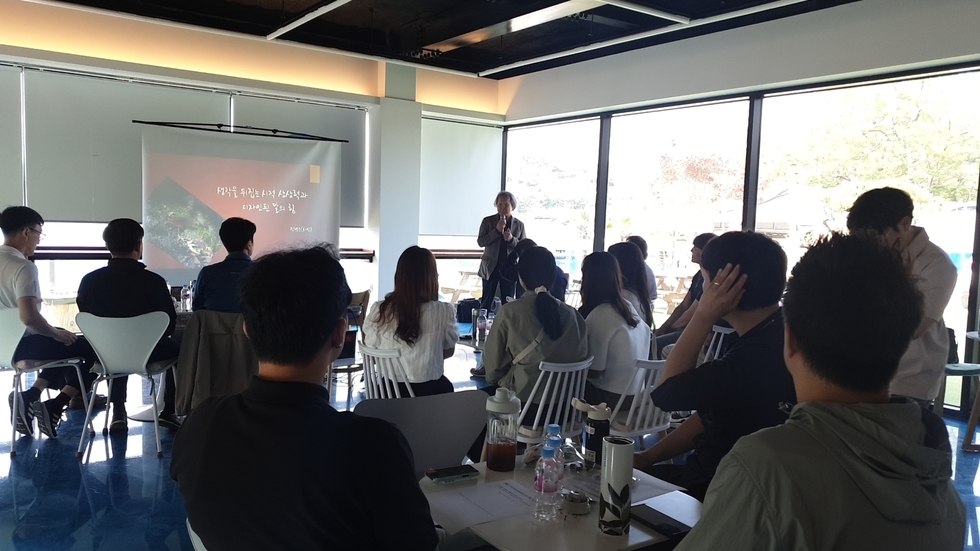 넓은 유리창이 있는 밝은 강의실에서 20여 명의 젊은 남녀들이 테이블에 앉아 한 강연자의 발표를 경청하고 있다. 강연자는 앞에 설치된 스크린 옆에 서서 마이크를 들고 있으며, 스크린에는 '생각을 뒤집는 시적 상상력과 디자인된 말의 힘'이라는 제목이 적혀 있다. 참석자들은 노트와 텀블러, 음료 등을 테이블 위에 올려놓고 집중하는 모습이다.