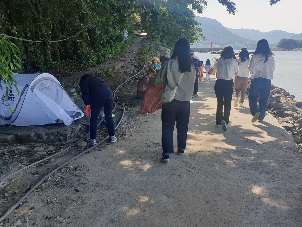 해변 산책로에서 여성들이 쓰레기봉투를 들고 해안가 정화활동을 진행하고 있다. 왼편에는 조그만 흰색 텐트가 펼쳐져 있고, 한 명은 장갑을 끼고 텐트 주변 쓰레기를 줍고 있다. 배경에는 나무 그늘과 바다가 어우러져 있으며, 멀리 빨간 등대와 산이 보인다. 참여자들은 대부분 2,30대이며 캐주얼한 옷차림이다.