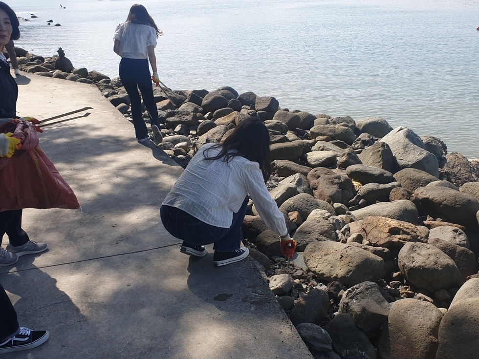 해변가 바위 사이에 버려진 쓰레기를 줍기 위해 여성 참가자가 쪼그려 앉아 집게로 정리하고 있다. 주변에는 함께 환경 정화활동에 참여 중인 사람들이 쓰레기봉투와 장갑을 착용한 채 해안가를 따라 걸으며 쓰레기를 수거하고 있다. 