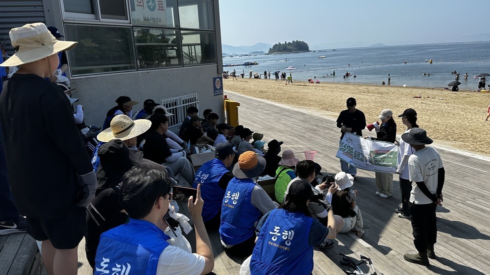 30여명의 사람들이 계단에 앉아있으며 캠페인을 진행하는 안내자의 안내 사항을 듣고 있는 모습이며 몇명은 휴대전화를 앞의 모습을 촬영하는 모습이다. 4명의 관계자들이 참여자들을 대상으로 설명중이다. 확성기를 든 여성과 플랜카드를 든 2명의 남성, 옆에는 이를 바라보고 있는 모습이다.  