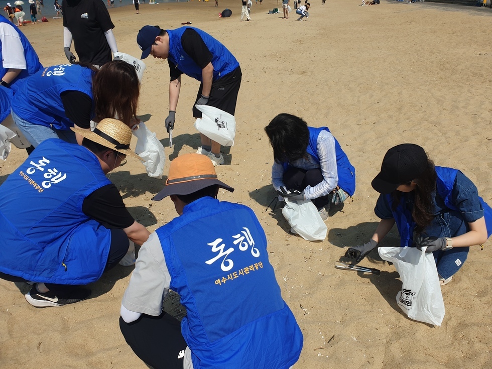 '동행 여수시도시관리공단' 이라는 문구가 적힌 파란색 조끼를 입은 8명의 남녀가 해변에서 쓰레기 수거 활동을 하고 있다. 몇 명은 바닥에 앉아 손에 비닐봉지를 들고 쓰레기를 주우며 활동에 참여하고 있다.