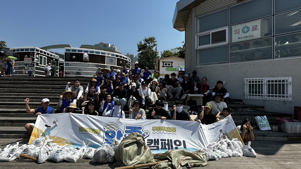 해변에서 진행된 '알줍캠페인' 캠페인 활동을 마친 후 단체 기념촬영을 하고 있다. 약 50명이 계단에 앉아 '알려주세요! 주워주세요! 알줍 캠페인' 이라는 문구가 적힌 현수막을 들고 포즈를 취하며 웃고 있다. 쓰레기봉투는 현수막 앞에 정리되어 있다.