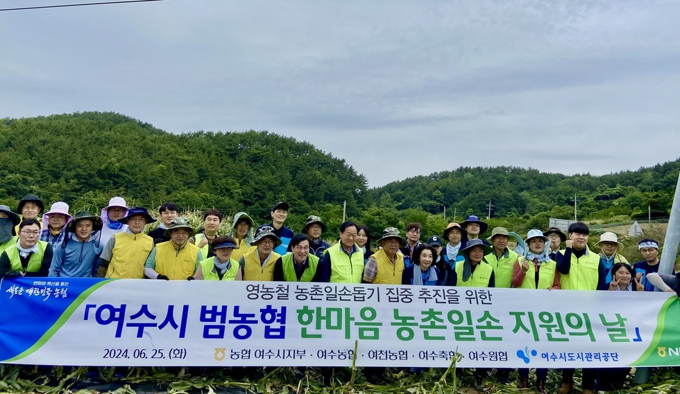 '영농철 농촌일손돕기 집중 추진을 위한 여수시 범농협 한마음 농촌일손 지원의 날 2024.06.25(화) 농협 여수시지부 여천농협 여수축협 여수원협 여수시도시관리공단' 이란느 문구가 쓰여진 현수막을 들고 단체사진을 촬영 중인 참가자들. 다양한 연령대의 50여명의 사람들이 모자를 쓰고 노란색, 연두색 조끼 등을 착용한 채 밝게 웃고 있으며, 뒤편에는 푸른 숲이 펼쳐져 있는 전형적인 농촌 풍경이 보인다.