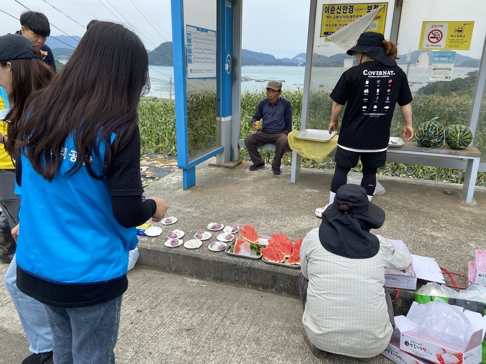 버스정류장 쉼터에서 수박과 떡 등 간식을 나누어 먹으며 잠시 휴식을 취하고 있는 모습. 정류장 벤치에는 남자 어르신 한 분이 앉아 음료를 마시고 있고, 다른 참가자들은 바닥에 놓인 접시를 정리하거나 간식을 준비하고 있다.