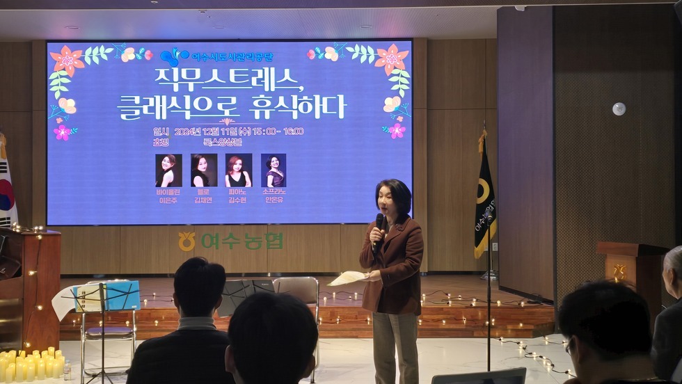 화면의 스크린에는 여수시도시관리공단 직무스트레스, 클래식으로 휴식하다 일시 2024년 12월 11일 (수) 15:00 ~ 16:00 쵸빙 룩스앙상블 바이올린 이은주 첼로 김채연 피아노 김수현 소프라노 안온유 라는 문구가 적혀있는 화면이 띄워져 있으며 50대 여성으로 보이는 여성이 마이크와 행사지를 들고 행사를 진행하고 있고, 임직원들은 경청하는 모습이다.