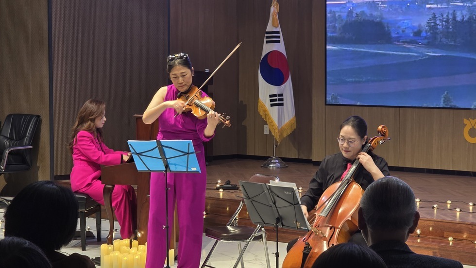무대 위에서 보라색 옷을 입은 여성이 바이올린을 연주하고 있고, 왼편에는 분홍색 정장을 입은 여성이 피아노를 연주하고 있으며, 오른편에는 검은색 옷을 입은 여성이 첼로를 연주하고 있다. 무대 뒤에는 태극기가 세워져 있고, 관객들이 연주를 감상하고 있는 모습이다.