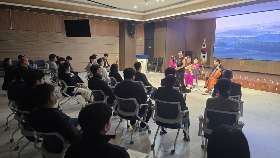 무대 위에서 검은색 드레스를 입은 여성이 노래를 부르고 있으며, 뒤에는 바이올린, 첼로, 피아노 연주자가 연주를 하고 있다. 객석에는 30여명의 여수시도시관리공단 임직원들이 앉아 진지하게 공연을 감상하고 있다. 무대 오른쪽에는 태극기가 세워져 있고, 배경 스크린에는 풍경 사진이 보인다.