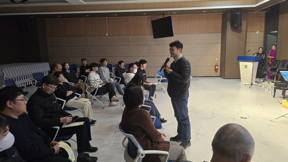 여수시도시관리공단 클래식 공연 행사 중 한 남성이 마이크를 들고 관객 앞에서 이야기하고 있다. 객석에는 30여명의 임직원들이 앉아 경청하고 있으며, 무대 오른쪽에는 연주자들이 대기 중이다. 배경에는 나무 패널로 마감된 벽면과 벽걸이 TV, 연주용 마이크가 세팅된 단상이 보인다.