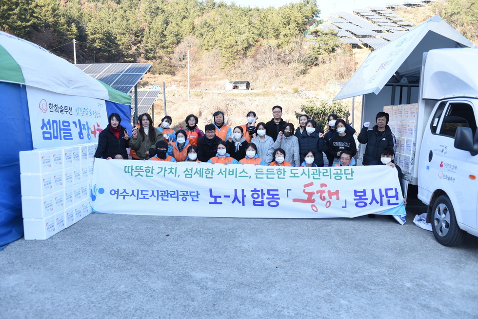 ‘따뜻한 가치, 섬세한 서비스, 든든한 도시관리공단 여수시도시관리공단 노·사 합동 동행 봉사단’이라는 문구가 적힌 현수막을 들고 여수시도시관리공단 임직원들이 김장 나눔 봉사활동을 기념하며 단체 사진을 촬영한 모습이다. 다양한 연령대의 임직원 20여 명이 모였으며, 일부는 오렌지색 앞치마와 마스크를 착용한 상태로, 김장박스가 가득 실린 트럭과 텐트 옆에서 밝은 표정으로 포즈를 취하고 있다. 