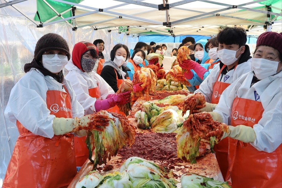 20여명의 참여자들이 김장을 하고 있는 모습이다. 하얀 방역복과 주황색 앞치마, 고무장갑을 착용한 참가자들은 김장 테이블을 따라 길게 줄지어 서 있으며, 배추 속을 정성스럽게 버무려가며 김치를 만드는 과정이 활기차게 진행되고 있다. 모두가 밝은 표정으로 김치를 들고 카메라를 향해 포즈를 취하고 있다.