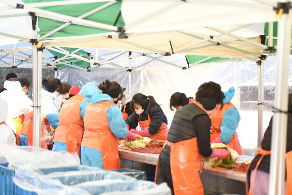 20여명의 참여자들이 김장을 하고 있는 모습이다. 하얀 방역복과 주황색 앞치마, 고무장갑을 착용한 참가자들은 김장 테이블을 따라 길게 줄지어 서 있으며, 배추 속을 정성스럽게 버무려가며 김치를 만드는 모습이다. 