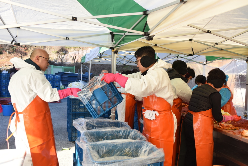 1김장 나눔 봉사 현장에서 한명의 남성 참여자가 완성된 김치를 담은 용기를 앞에 있는 남성 참여자에게 전달해주고 있는 장면이다. 뒷편에는 10여명의 사람들이 김장을 진행하고 있는 모습이다. 