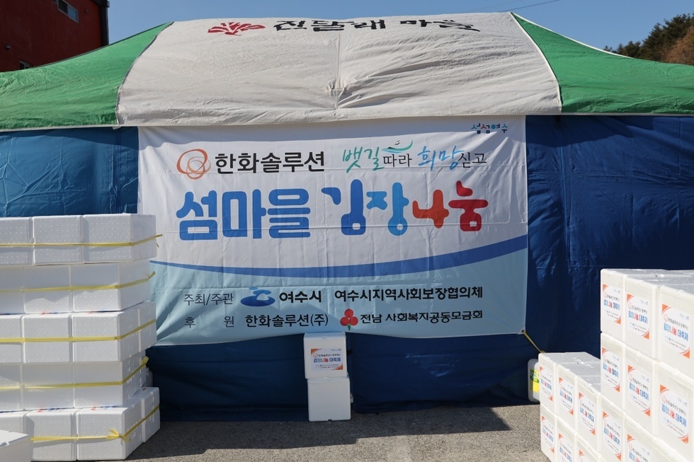 천막에는 '한화솔루션 뱃길 따라 희망 싣고 섬마을 김장나눔 '이라는 문구가 적혀 있으며, 주최/주관은 여수시, 여수시지역사회보장협의체, 후원은 한화솔루션(주), 전남 사회복지공동모금회로 표시되어 있다.  주변에는 다수의 하얀색 김장용 스티로폼 박스들이 정돈되어 쌓여 있는 모습이다. 