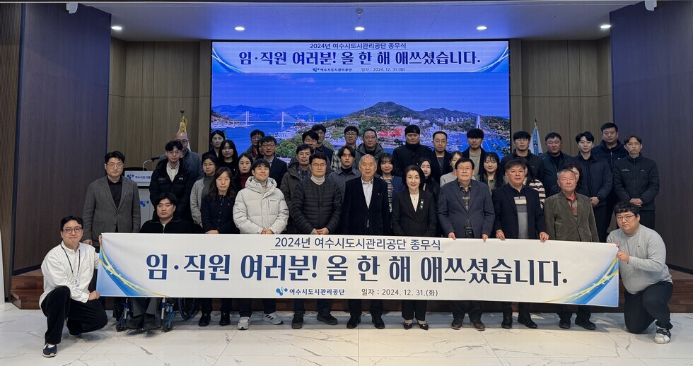 '2024년 여수시도시관리공단 종무식 임·직원 여러분! 올 한 해 애쓰셨습니다. 여수시도시관리공단 일자 : 2024.21.31 (화)'라는 문구가 적힌 대형 화면을 배경으로 같은 문구가 적혀진 현수막을 들고 여수시도시관리공단 임직원들이 단체로 촬영한 기념사진이다. 사진에는 30여 명의 임직원이 참여했으며 연령대는 20대에서 60대까지 다양하다. 2명의 남자 직원의 현수막을 길에 펼치며 앉아 있고, 가운데에는 50대 남성과 여성이 나란히 서 있으며, 전체적으로 단정한 복장으로 정돈된 분위기 속에서 한 해의 마무리를 기념하고 있다.