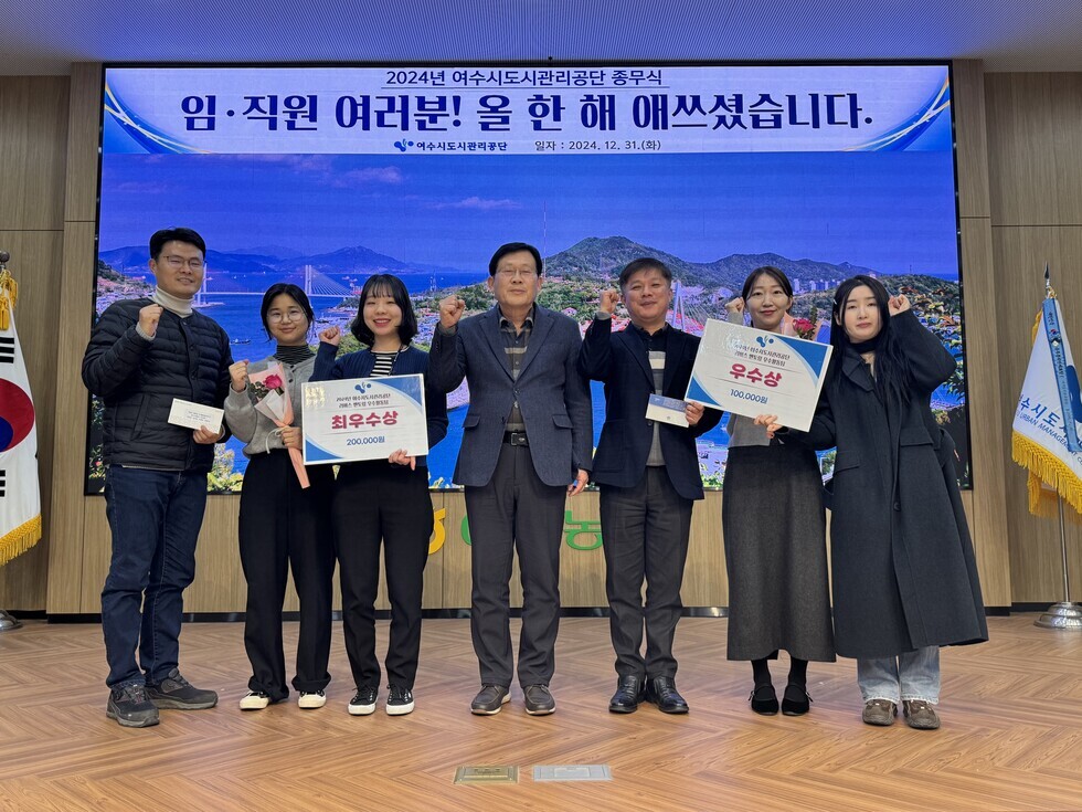 '2024년 여수시도시관리공단 종무식 임·직원 여러분! 올 한 해 애쓰셨습니다. 여수시도시관리공단 일자 : 2024.21.31 (화)'라는 문구가 적힌 대형 화면을 배경으로  7명의 임직원들이 포즈를 취하며 사진을 찍고 있다.  '최우수상'과 '우수상' 수상자들이 상장과 함께 꽃다발 및 상품권을 들고 있으며, 함께 기념 촬영한 임직원들은 20대부터 50대까지의 연령대로 구성되어 있다. 참여자들은 모두 환한 미소와 주먹 인사 포즈로 밝은 분위기를 연출하고 있다.