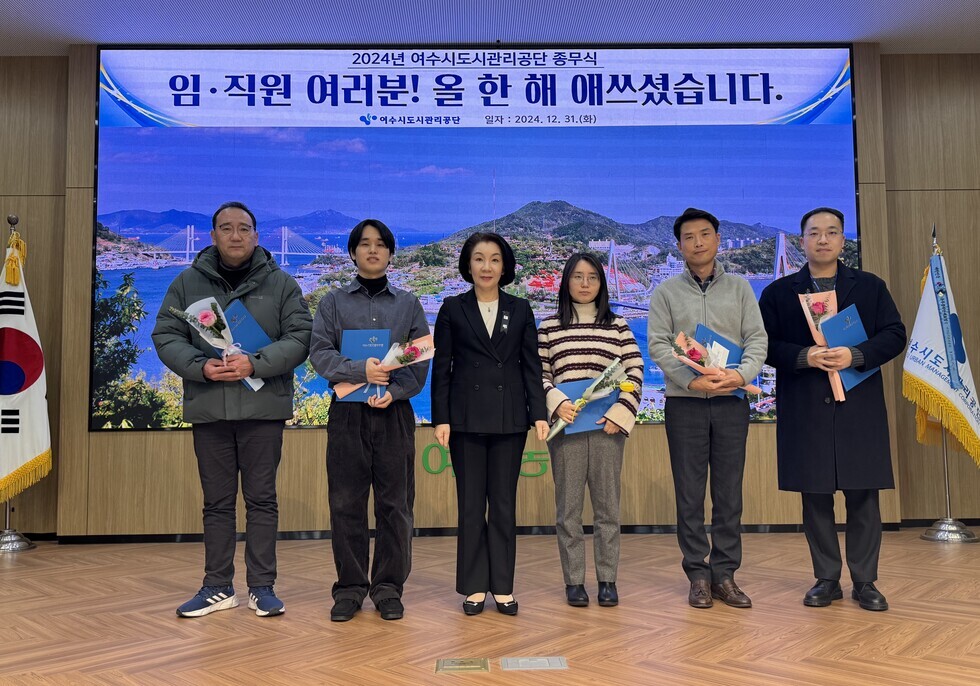 '2024년 여수시도시관리공단 종무식 임·직원 여러분! 올 한 해 애쓰셨습니다. 여수시도시관리공단 일자 : 2024.21.31 (화)'라는 문구가 적힌 대형 화면을 배경으로  6명의 임직원들이 포즈를 취하며 사진을 찍고 있다.  상장과 함께 꽃다발 및 상품권을 들고 있으며, 함께 기념 촬영한 임직원들은 20대부터 50대까지의 연령대로 구성되어 있다. 