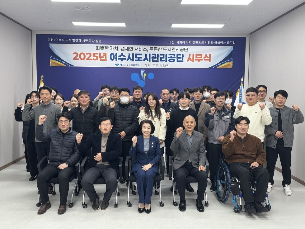 '따뜻한 가치, 섬세한 서비스, 든든한 도시관리공단 2025년 여수시도시관리공단 시무식 여수도시관리공단 일자 : 2025.1.2(목)'이라는 문구가 적힌 현수막을 배경으로, 여수시도시관리공단 임직원들이 시무식을 기념하여 단체 촬영한 사진이다. 총 30여 명의 임직원이 참여했으며 연령대는 20대부터 60대까지 다양하다. 앞줄에는 4명이 의자에 앉아 있고, 오른쪽에는 휠체어를 탄 인물이 위치해 있다. 그 뒤편으로는 여러 명의 임직원들이 힘찬 주먹 인사를 하며 포즈를 취하고 있다.