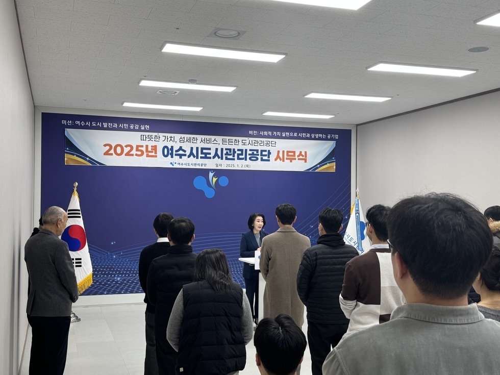 '따뜻한 가치, 섬세한 서비스, 든든한 도시관리공단 2025년 여수시도시관리공단 시무식 여수시도시관리공단 일자: 2025.1.2(목)'라는 문구가 적힌 현수막이 걸려 있는 실내 공간에서 여수시도시관리공단 시무식이 진행 중인 장면이다. 중앙 연단에는 정장을 입은 여성(50대 추정)이 마이크를 들고 발언을 하고 있으며, 그 앞에는 20대부터 60대까지 다양한 연령대의 임직원들이 단정하게 서서 경청하고 있다. 좌측에는 태극기가, 우측에는 공단기를 함께 배치하여 공식적인 분위기를 조성하고 있다.