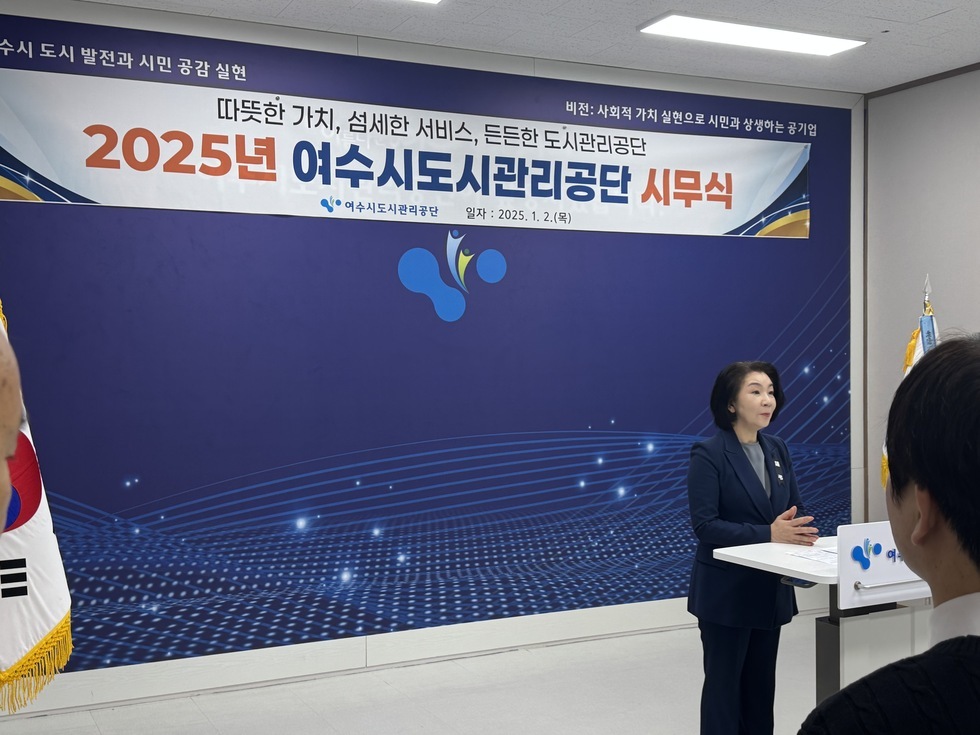 '따뜻한 가치, 섬세한 서비스, 든든한 도시관리공단 2025년 여수시도시관리공단 시무식 여수시도시관리공단 일자: 2025.1.2(목)'이라는 문구가 적힌 현수막이 걸린 실내에서 시무식이 진행 중이다. 연단 앞에는 정장을 입은 50대 여성 임직원이 서서 발언을 하고 있으며, 그 앞에는 여러 임직원들이 서서 경청하고 있는 모습이다. 좌측에는 태극기가, 우측에는 공단기가 배치되어 있어 공적인 분위기를 강조하고 있다.