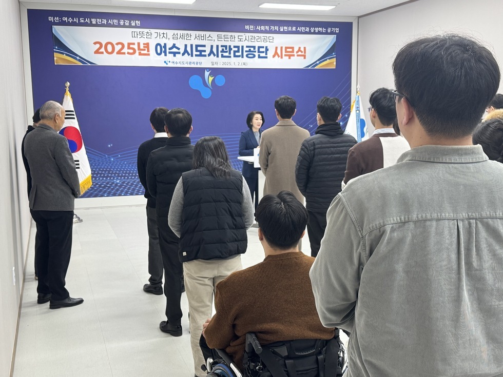 '따뜻한 가치, 섬세한 서비스, 든든한 도시관리공단 2025년 여수시도시관리공단 시무식 여수시도시관리공단 일자: 2025.1.2(목)'라는 문구가 적힌 현수막이 걸려 있는 실내 공간에서 여수시도시관리공단 시무식이 진행 중인 장면이다. 중앙 연단에는 정장을 입은 여성(50대 추정)이 마이크를 들고 발언을 하고 있으며, 그 앞에는 20대부터 60대까지 다양한 연령대의 임직원들이 단정하게 서서 경청하고 있다. 좌측에는 태극기가, 우측에는 공단기를 함께 배치하여 공식적인 분위기를 조성하고 있다.
