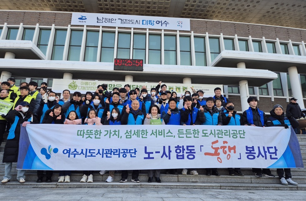 '따뜻한 가치, 섬세한 서비스, 든든한 도시관리공단 여수시도시관리공단 노‧사 합동 동행 봉사단' 이라는 문구가 적힌 대형 현수막을 펼쳐 들고, 그 뒤로는 '아름다운 나눔보따리 희망을 배달합니다.' 등의 문구가 적힌 현수막이 붙은 여수시청 앞 계단에서 기념촬영한 모습이다. 여수 시청 건물에는 여수시 남해안 거점도시 미항 여수라는 현수막이 걸려있다. 생필품이 담긴 나눔 보따리를 전달하기 위해 모인 여수시도시관리공단 봉사단 50여 명이 단체로 서 있으며, 대체로 20대에서 50대 연령대의 직원들로 구성되어 있다. 일부는 손가락 하트나 엄지손가락을 들어 보이며 포즈를 취하고 있다.