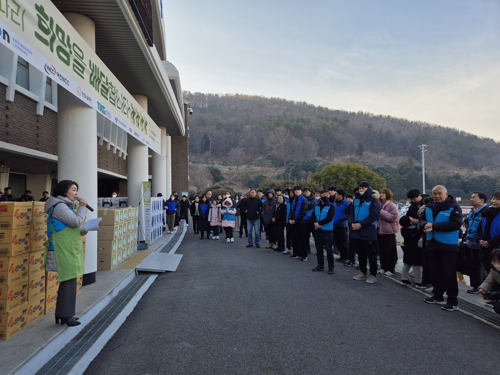 '아름다운 보따리 희망을 배달합니다' 라는 문구가 적힌 현수막이 걸려 있는 건물 앞에서 여수시도시관리공단 여자 관계자가 마이크를 들고 행사 안내문을 읽고 있으며, 그 뒤에는 생필품 박스들이 정리되어 쌓여 있다. 행사에 참여한 50여 명의 봉사자들이 넓게 줄지어 서서 안내를 경청하고 있으며, 대부분 파란색 단체 점퍼를 착용한 20대에서 60대 연령대의 시민과 임직원들로 구성되어 있다.