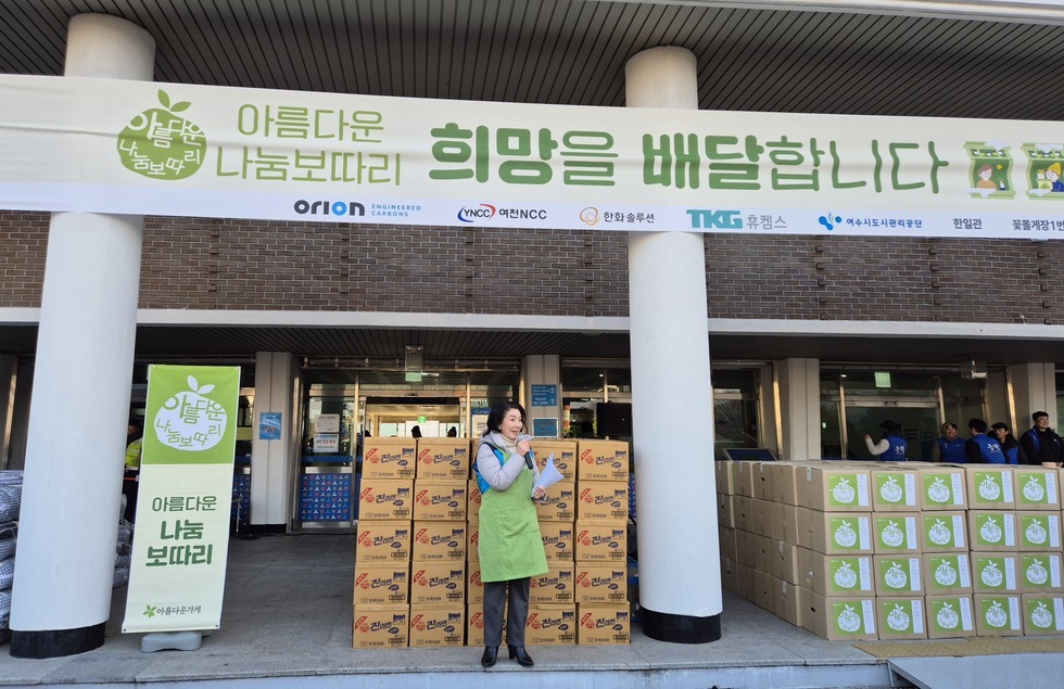 '아름다운 나눔보따리 희망을 배달합니다 (하단 문구 : ORION, YNCC 여천NCC, 한화솔루션, 휴켐스,여수시도시관리공단 한일관, 꽃돌게장)' 라는 문구가 적힌 대형 현수막이 걸린 건물 앞에서, 초록색 앞치마를 착용한 여성이 마이크와 종이를 들고 행사를 진행하고 있다. 여성 뒤로는 '아름다운 나눔보따리' 로고가 인쇄된 박스들과 '진라면' 상자들이 높게 쌓여 있으며, 현장 좌측에는 '아름다운 나눔 보따리' 문구가 적힌 세로형 배너도 세워져 있다. 건물 안쪽에는 파란 조끼를 입은 행사 관계자들이 준비를 하고 있는 모습이 보인다.