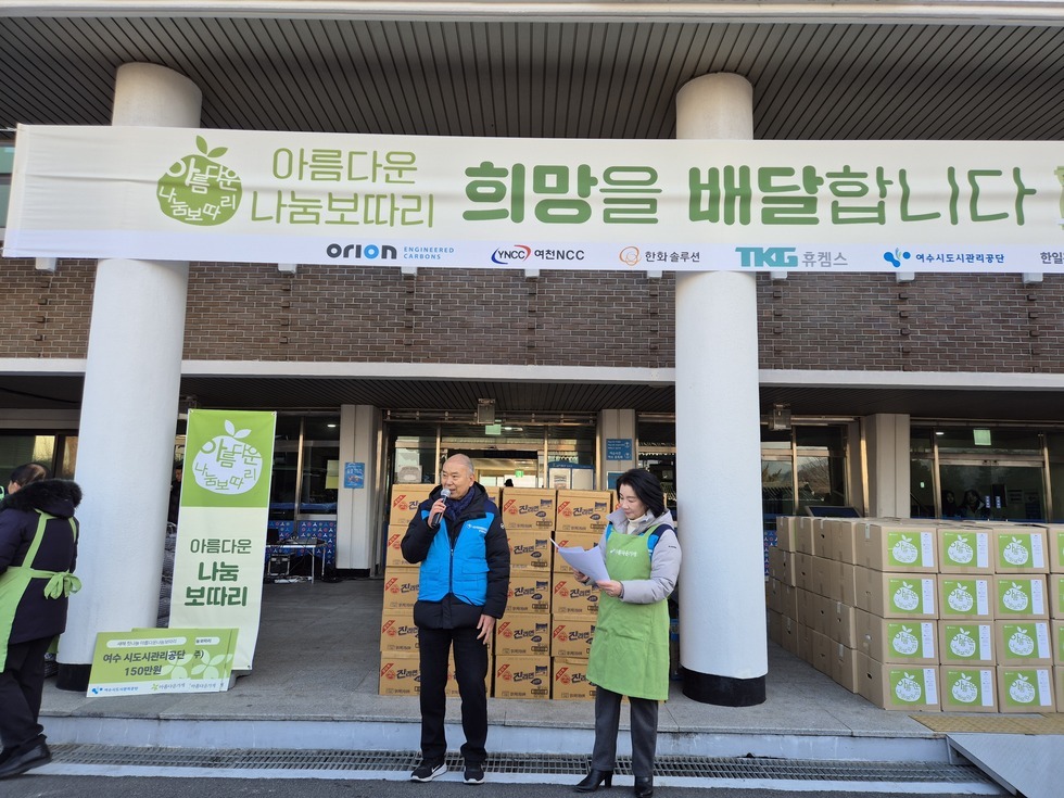 '아름다운 나눔보따리 희망을 배달합니다 (하단 문구 : ORION, YNCC 여천NCC, 한화솔루션, 휴켐스,여수시도시관리공단 한일관, 꽃돌게장)' 라는 문구가 적힌 대형 현수막이 걸린 건물 앞에서 여수시도시관리공단 관계자 두 명이 마이크와 자료를 들고 행사를 진행하고 있다. 60대 남성은 파란색 패딩 조끼를 입고 마이크를 잡고 있으며, 40대 여성은 초록색 앞치마를 두르고 종이를 읽고 있다. 뒤편에는 '진라면' 박스와 '아름다운 나눔보따리' 로고가 인쇄된 박스들이 높게 정리되어 있고, 좌측에는 '여수시도시관리공단 150만원 후원' 문구가 적힌 팻말과 '다름다운 나눔 보따리' 라는 문구가 적힌 세로형 배너가 함께 설치되어 있다.