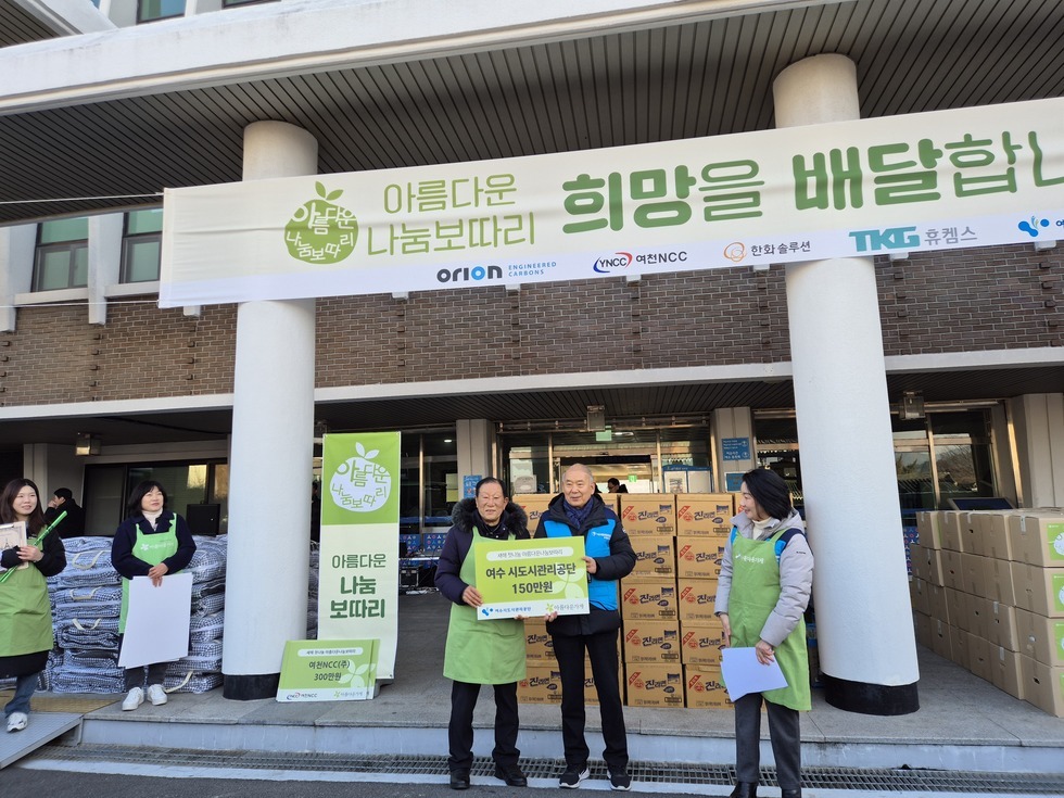 '아름다운 나눔보따리 희망을 배달합니다 (하단 문구 : ORION, YNCC 여천NCC, 한화솔루션, 휴켐스,여수시도시관리공단 한일관, 꽃돌게장)' 라는 문구가 적힌 대형 현수막 아래, '여수시도시관리공단 150만원' 이라고 적힌 기부판을 양손으로 들고 있는 60대 남성 두 사람이 나란히 서 있고, 초록색 앞치마를 착용한 행사 관계자들이 양옆에서 지켜보며 미소 짓고 있다. 그 뒤로는 '진라면' 박스와 '아름다운 나눔보따리' 로고가 붙은 박스들이 정돈되어 있으며, 왼쪽에는 '아름다운 나눔보따리', '여천NCC(주) 300만원'이라는 문구가 적힌 펫말과 '아름다운 나눔 보따리 '라는 문구가 적힌 세로형 배너와 팻말이 함께 세워져 있다.