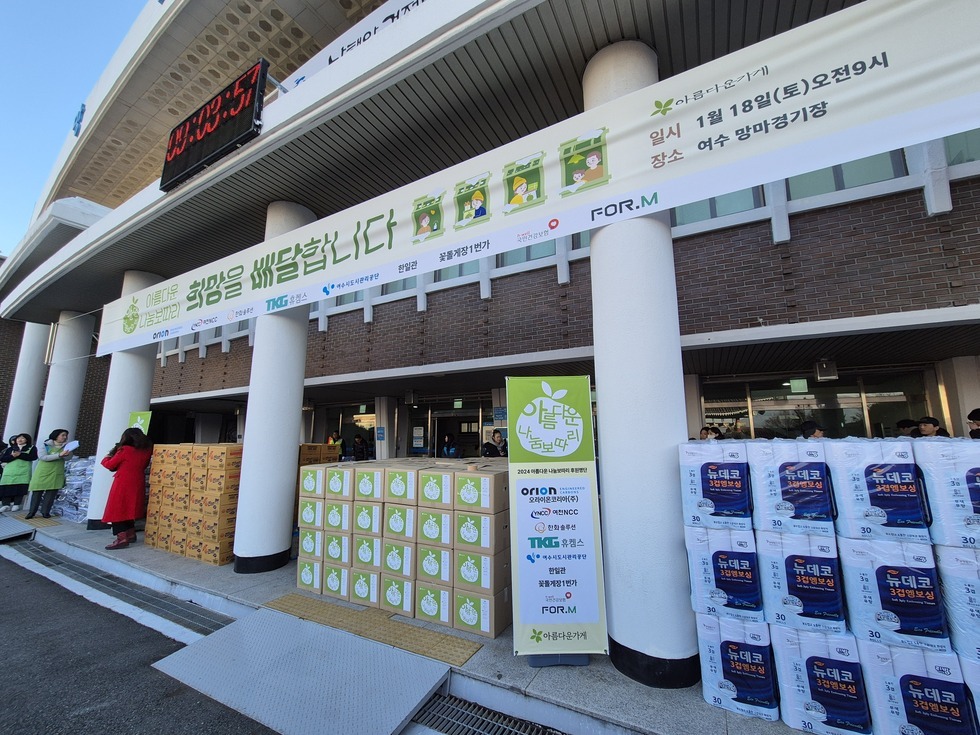 '아름다운 나눔보따리 희망을 배달합니다 일시 1월 18일(토) 오전 9시 장소 여수 망마경기장 ' 라는 문구가 적힌 대형 현수막이 건물 입구 상단에 길게 걸려 있고, 행사장 앞에는 '아름다운 나눔보따리' 로고가 붙은 박스들과 '뉴데코' 화장지 박스가 깔끔하게 정렬돼 있으며, 초록색 앞치마를 입은 행사 관계자들이 배분 준비를 하고 있다. 중앙에는 후원기관 로고와 명단이 적힌 세로 배너가 설치되어 있다.