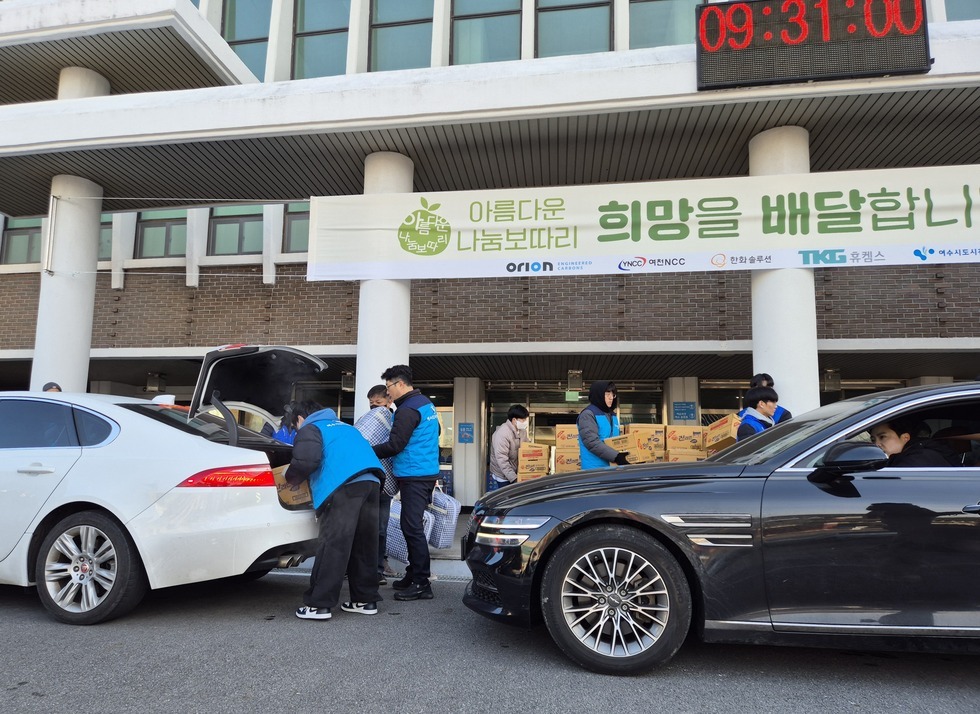 '아름다운 나눔보따리 희망을 배달합니다' 라는 문구가 적힌 현수막이 걸린 건물 앞에서, 파란 조끼를 착용한 봉사자들이 하얀색 차량의 트렁크에 생필품 상자와 담요 등을 차곡차곡 실어 나르고 있다. 검정 차량과 흰색 차량이 나란히 정차해 있으며, 검정 차량 안에는 남자 운전자가 대기 중이다. 건물 앞에는 '진라면' 박스들이 쌓여 있고, 그 앞에서 봉사자들이 활발히 물품을 분배하고 있다. 상단 전광판에는 '09:31:00'이라는 시간이 표시되어 있다.