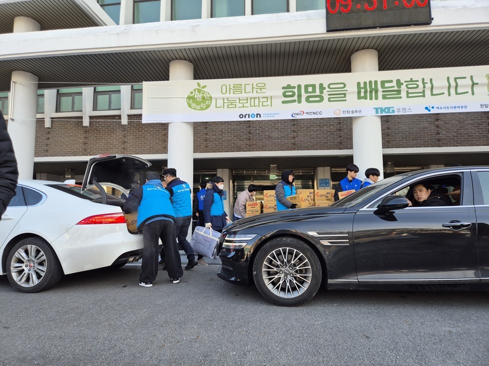 '아름다운 나눔보따리 희망을 배달합니다' 라는 문구가 적힌 현수막이 걸린 건물 앞에서, 파란 조끼를 착용한 봉사자들이 하얀색 차량의 트렁크에 생필품 상자와 담요 등을 차곡차곡 실어 나르고 있다. 검정 차량과 흰색 차량이 나란히 정차해 있으며, 검정 차량 안에는 남자 운전자가 대기 중이다. 건물 앞에는 '진라면' 박스들이 쌓여 있고, 그 앞에서 봉사자들이 활발히 물품을 분배하고 있다. 상단 전광판에는 '09:31:00'이라는 시간이 표시되어 있다.