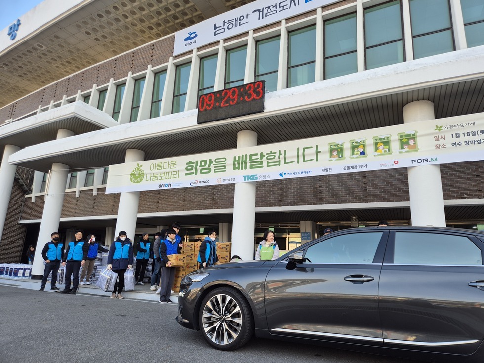 아름다운 나눔보따리 희망을 배달합니다' 라는 문구가 적힌 대형 현수막이 건물 전면에 걸려 있고, 전광판에는 '09:29:33'이라는 시간이 표시돼 있다. 행사 관계자들이 파란색 조끼와 초록 앞치마를 착용한 채 박스를 나르고 있으며, 생필품이 담긴 나눔보따리를 차량에 실을 준비를 하고 있다. 건물 앞에는 검정 승용차가 정차해 있고, 주변에는 담요와 라면 박스가 정돈되어 쌓여 있다.