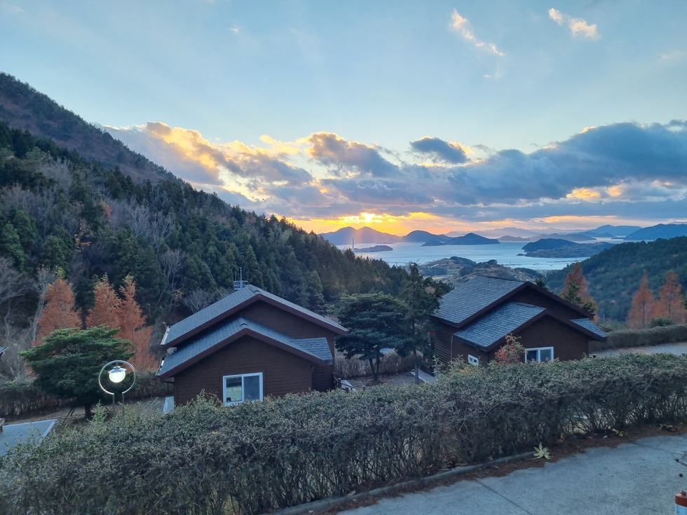 해가 뜨는 하늘 아래로 섬과 바다가 어우러진 풍경이 펼쳐지고, 그 앞에 봉황산자연휴양림의 갈색 목조 숙소 두 채가 자리하고 있다. 해가 산 너머로 떨어지며 붉은빛과 푸른빛이 교차하는 하늘과 구름이 장관을 이루고, 숲으로 둘러싸인 숙소 주변은 조용하고 평화로운 분위기를 자아낸다. 해송과 낙엽송이 어우러진 자연경관 속에서 머무르며 일몰을 감상할 수 있는 포근한 휴식처의 모습이다.