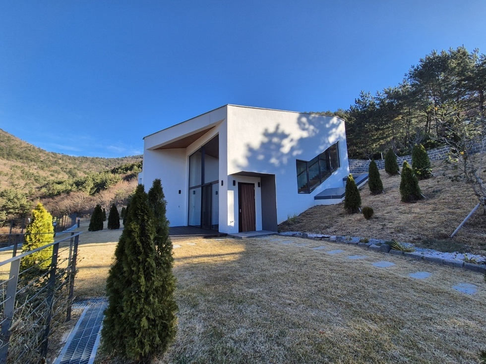 푸른 하늘 아래, 봉황산자연휴양림에 위치한 모던 스타일의 신축 건물이 깔끔하게 들어서 있다. 새하얀 외벽과 비대칭 구조의 지붕, 전면 유리창이 어우러져 세련된 분위기를 자아내며, 단정하게 다듬어진 잔디밭과 소나무형 조경수들이 주변을 감싸고 있다. 건물 옆으로는 계단식 석축이 이어지며 산비탈을 따라 자연스럽게 조성된 정원과 어우러진다. 현대적인 건축미가 돋보이는 이 건물은 자연 속에서도 도심의 감성을 느낄 수 있는 휴양공간으로 보인다.