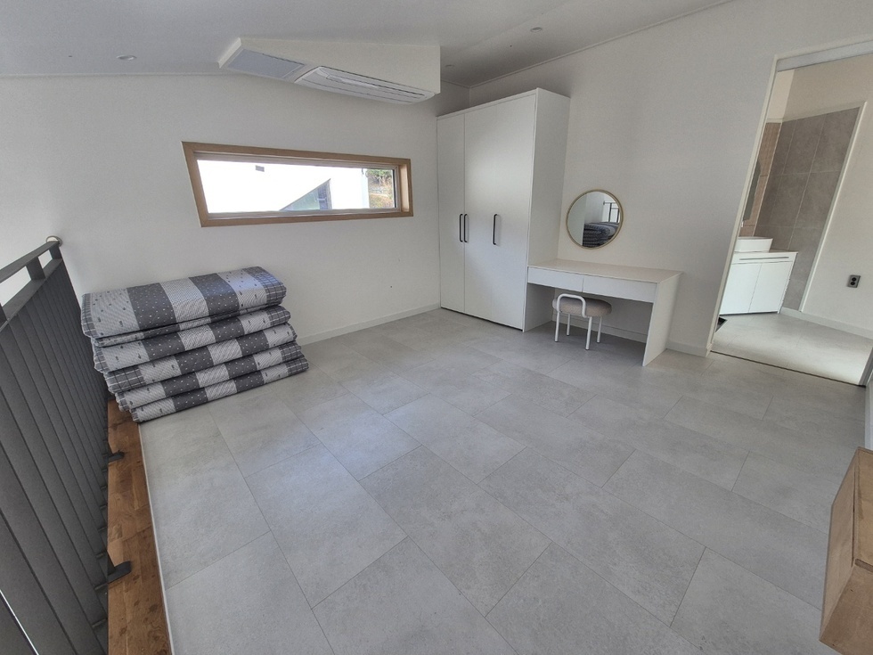 봉황산자연휴양림 신축동의 실내 모습으로, 밝고 깔끔한 분위기의 침실 공간이다. 바닥은 은은한 회색 타일로 마감되어 있으며, 왼쪽에는 접이식 요와 이불이 가지런히 쌓여 있고, 오른쪽에는 흰색 옷장과 원형 거울이 놓인 화장대, 의자가 배치되어 있다. 창문은 가로로 길게 나 있어 채광이 좋고, 창 너머로 외부 풍경도 일부 보인다. 오른쪽 문 너머에는 밝은 톤의 타일로 마감된 욕실이 연결되어 있으며, 전반적으로 심플하면서도 기능적인 구조를 갖춘 공간이다.