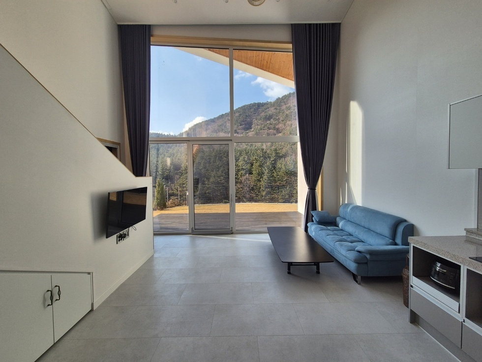 봉황산자연휴양림 신축동의 거실 내부 모습으로, 천장이 높고 전면이 통유리로 되어 있어 개방감이 뛰어나다. 유리창 너머로는 숲과 산이 어우러진 자연 경관이 시원하게 펼쳐지며, 실내로 풍부한 자연광이 들어온다. 오른쪽에는 짙은 블루 컬러의 3인용 소파와 낮은 원목 테이블이 놓여 있어 편안한 휴식 공간을 제공한다. 왼편에는 벽걸이 TV가 설치되어 있고, 아래쪽에는 수납장이 배치되어 있다. 심플하고 현대적인 인테리어가 자연 속에서의 휴식을 더욱 쾌적하게 만들어준다.