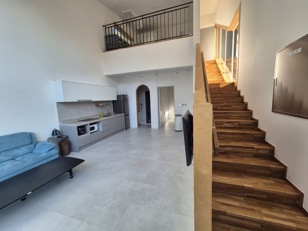 모던한 구조의 복층형 숙소 내부를 촬영한 이미지입니다. 오른쪽에는 원목 계단이 있고, 계단 옆으로는 큰 창을 통해 자연광이 들어오고 있습니다. 왼쪽에는 블루 컬러의 소파와 낮은 테이블, 그리고 일체형 싱크대와 전자레인지, 냉장고 등이 갖춰진 주방 공간이 있으며, 중간 위쪽에는 복층 공간이 보입니다. 전체적으로 화이트 톤의 벽과 바닥 타일로 깔끔하고 현대적인 느낌을 줍니다.