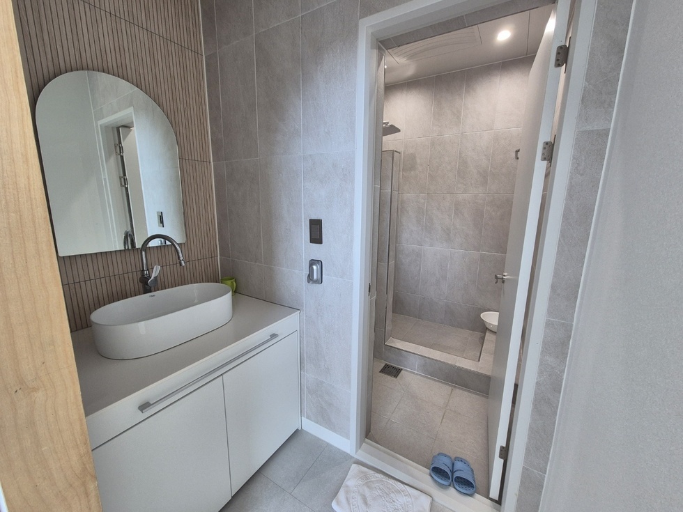 봉황산자연휴양림 신축동의 욕실 내부 모습으로, 세면대와 샤워실이 분리된 구조로 깔끔하게 구성되어 있다. 전반적으로 회색 타일을 사용하여 차분하고 모던한 분위기를 자아내며, 세면대는 넓은 상판 위에 원형 라인의 세련된 화이트 세면볼이 놓여 있고, 그 위로는 아치형 거울이 설치되어 있다. 우측으로는 투명 유리 칸막이로 분리된 샤워 공간이 있으며, 안쪽에는 변기와 샤워기가 설치되어 있다. 욕실 입구 바닥에는 파란 슬리퍼가 가지런히 놓여 있어 실용적인 면도 더해준다.