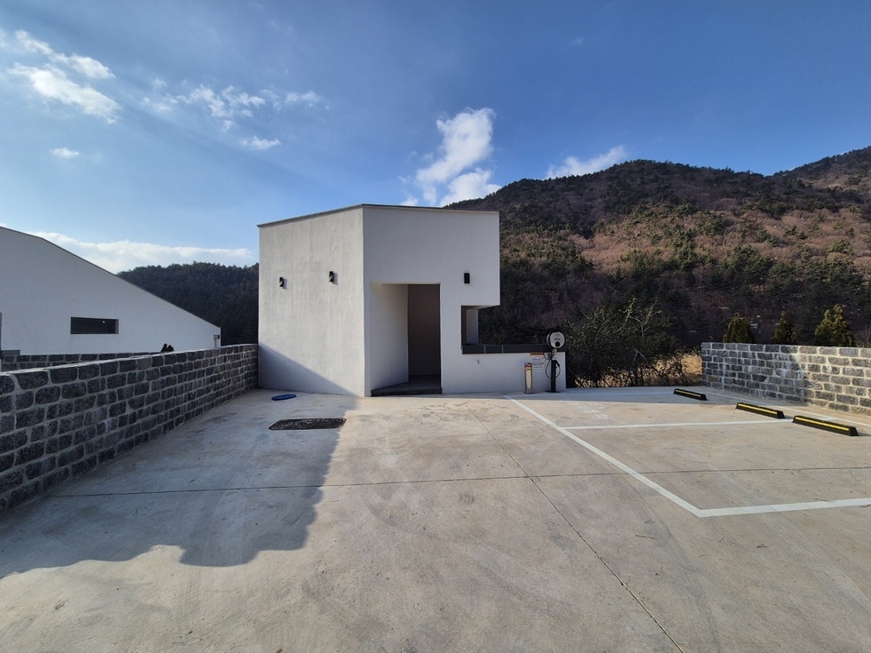 봉황산자연휴양림 신축동 외부 주차장 전경으로, 흰색 외벽의 미니멀한 건축물과 함께 산자락이 배경을 이루고 있다. 건물 앞 넓은 콘크리트 바닥에는 주차구역이 정리되어 있으며, 주차라인과 차량 멈춤턱이 명확하게 표시되어 있다. 건물 오른편에는 전기차 충전기로 보이는 기기와 소화기가 설치되어 있어 안전과 친환경 요소도 고려한 공간임을 보여준다. 건물 좌측에는 낮은 석축 담장이 둘러져 있어 주변 자연 환경과 조화를 이루는 안정적인 구조를 형성하고 있다.