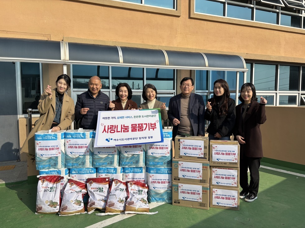 '따뜻한 가치, 섬세한 서비스, 든든한 도시관리공단 사랑나눔 물품기부 여수시도시관리공단 임직원 일동' 이라는 문구가 적힌 팻말을 들고 있고, '사랑나눔 물품기부' 라는 스티커가 붙은 쌀, 화장지, 생필품 박스 등이 정돈된 상태로 쌓여 있다. 해당 물품들을 기부한 여수시도시관리공단 임직원 7명(30대에서 50대 연령대)이 건물 외부 공간에서 손가락 하트 포즈를 취하며 기념촬영한 단체 사진이다.