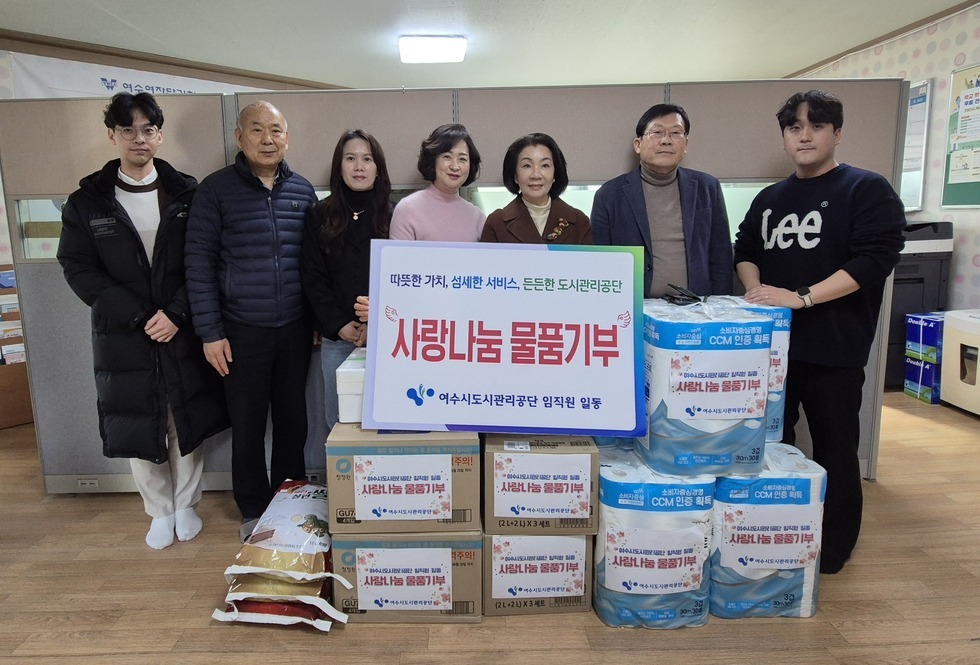 '따뜻한 가치, 섬세한 서비스, 든든한 도시관리공단 사랑나눔 물품기부 여수시도시관리공단 임직원 일동' 이라는 문구가 적힌 팻말을 들고 있고, 그 주변으로는 '사랑나눔 물품기부' 라는 스티커가 부착된 쌀, 화장지, 생필품 박스 등이 깔끔하게 정리되어 있다. 해당 물품들을 기부한 여수시도시관리공단 임직원 7명(20대에서 50대 연령대)이 실내 공간에서 팻말을 중심으로 나란히 서서 기념촬영한 단체 사진이다.