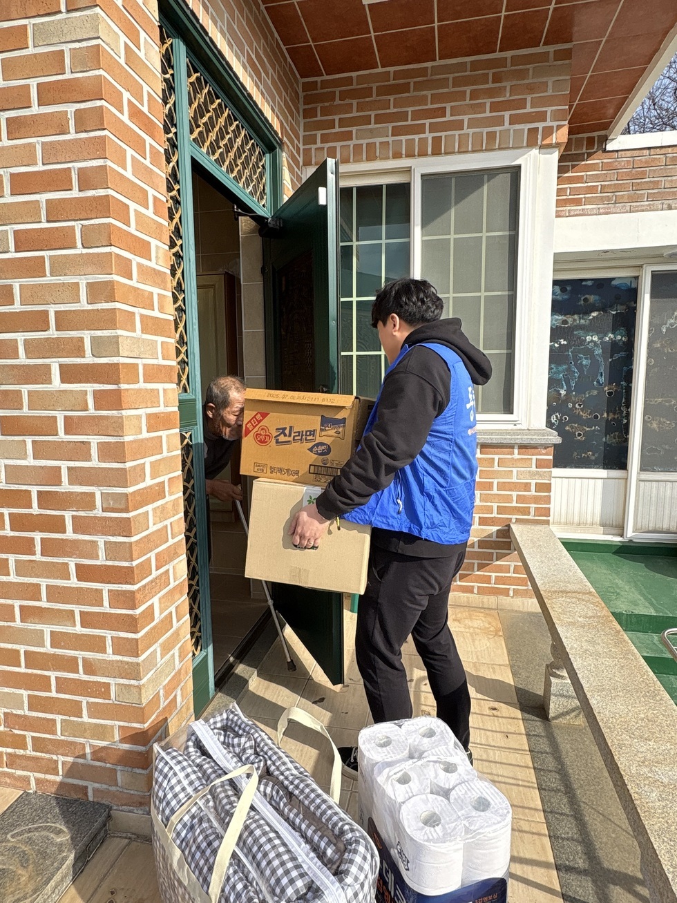 파란 조끼를 입은 20대 남성 봉사자가 벽돌 주택 현관 앞에서 '진라면' 박스와 생필품 상자가 담긴 상자를 들고 어르신에게 전달하고 있다. 문 안쪽에서는 머리가 희끗한 어르신이 문을 열어주고 있는 모습이며, 문 앞 바닥에는 담요 가방과 휴지 묶음 등 나눔보따리 물품이 놓여 있다. 겨울 햇살이 따스하게 비치는 가운데, 봉사자는 조심스럽게 물품을 전달하고 있다.