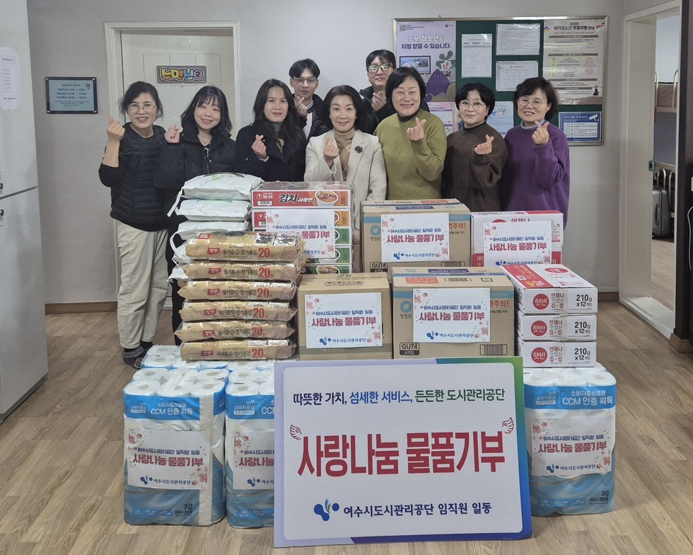 '따뜻한 가치, 섬세한 서비스, 든든한 도시관리공단 사랑나눔 물품기부 여수시도시관리공단 임직원 일동' 이라는 문구가 적힌 팻말이 세워져 있고, 그 뒤로는 '사랑나눔 물품기부' 라는 스티커가 붙은 쌀, 라면, 휴지, 생필품 박스 등이 정돈된 상태로 쌓여 있다. 해당 물품들을 기부한 여수시도시관리공단 이사장 및 임직원 9명이 물품 뒤에 서서 손가락 하트 포즈를 취하며 기념 촬영한 단체 사진이다.