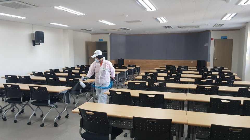 밝고 정돈된 강의실에서 방역 작업자가 연무형 소독기를 이용해 책상 사이를 소독하고 있다. 작업자는 흰색 보호장구와 마스크, 모자를 착용하고 있으며, 책상과 의자가 규칙적으로 배치된 공간에서 꼼꼼하게 방역을 수행 중이다. 천장에는 형광등이 켜져 있고, 전체적으로 청결하고 조용한 분위기다.