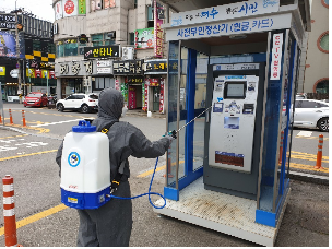 도심 거리에서 방역복을 착용한 작업자가 ATM기를 소독하고 있다. 작업자는 등짐형 분무기를 메고 있으며, 파란색 부스 안에 설치된 무인 ATM기를 향해 소독제를 분사하고 있다. 