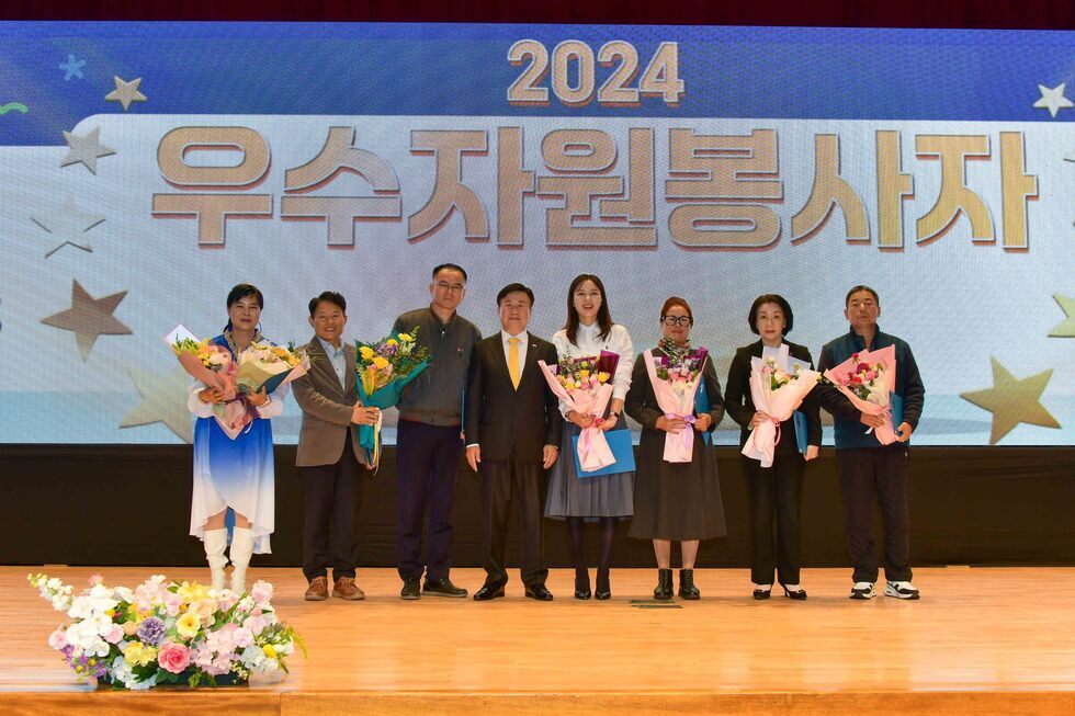'2024 우수자원봉사자' 라는 문구가 배경 전광판에 크게 보이는 무대 위에서, 수상자 8명이 꽃다발을 들고 기념촬영을 하고 있다. 가운데 정장을 입은 남성과 함께 양옆으로 각기 다른 의상을 입은 여성과 남성들이 서 있으며, 모두 밝은 표정으로 정면을 바라보고 있다. 무대 앞에는 꽃장식이 놓여 있고, 전체적으로 축하 분위기가 느껴진다. 연령대는 30대~ 60대이다. 