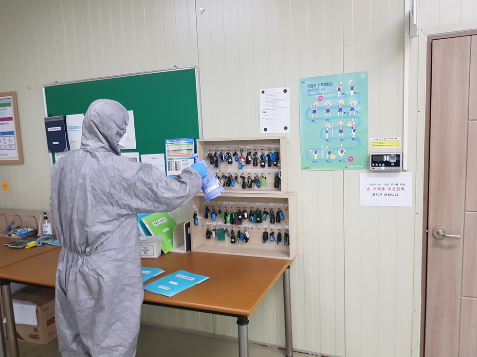 회색 방역복을 입은 남성 작업자가 사무실 내부에서 열쇠 보관함에 소독제를 분사하며 방역 작업을 진행하고 있다. 벽에 부착된 보관함에는 다양한 색상의 열쇠들이 걸려 있다.